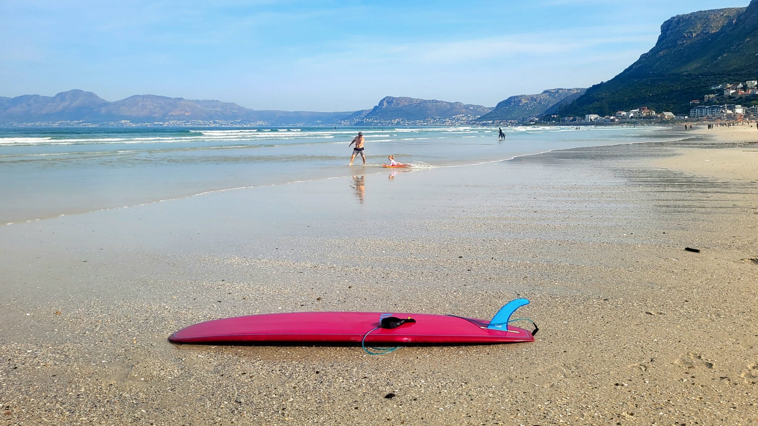 Pink surfboard on Muizenberg sandy beach with False Bay and mountains in the background